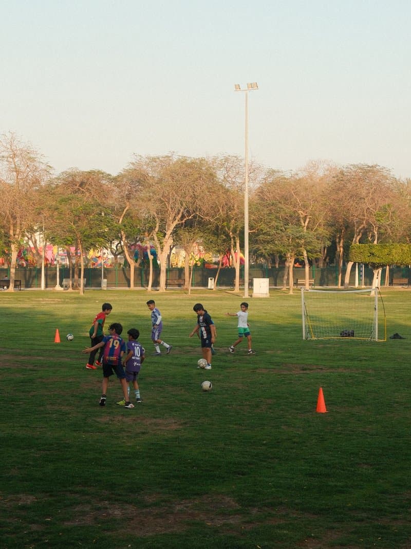 Children playing soccer on a grassy field with goalposts and trees in the background.