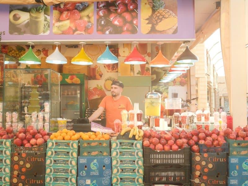 A fruit vendor stands behind a colorful display of fruits in a market, with vibrant hanging lights above.