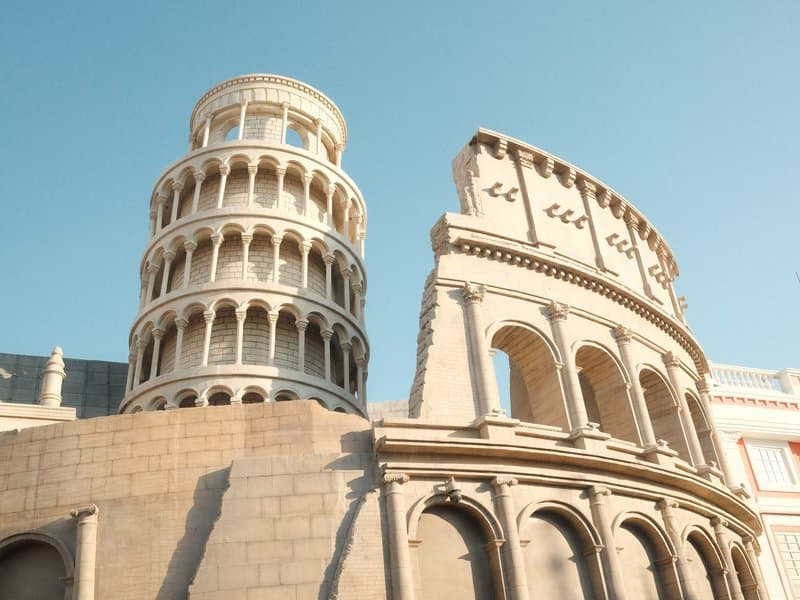 A close-up view of the Leaning Tower of Pisa and a Roman-style coliseum against a clear blue sky.