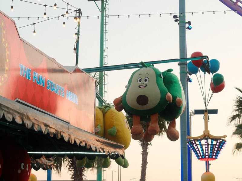 Colorful carnival scene featuring plush avocado and lemon toys hanging from a booth with festive lights.