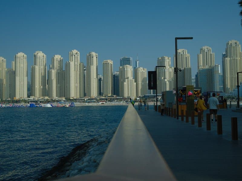 A waterfront promenade lined with tall buildings and people walking, under a clear blue sky.