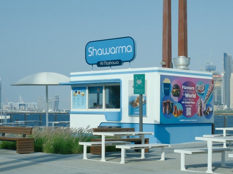 A colorful shawarma food kiosk by the waterfront with benches and a clear sky.
