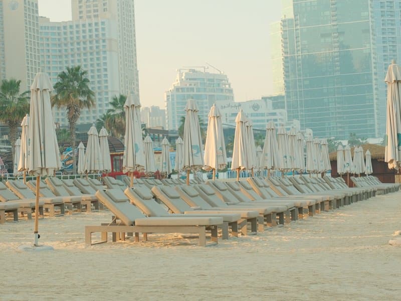 A beach scene with rows of sun loungers and umbrellas, surrounded by tall buildings in the background.