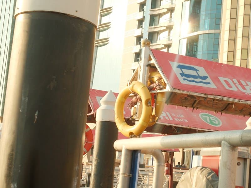 A yellow life ring hanging on a dock with modern buildings in the background under warm sunlight.