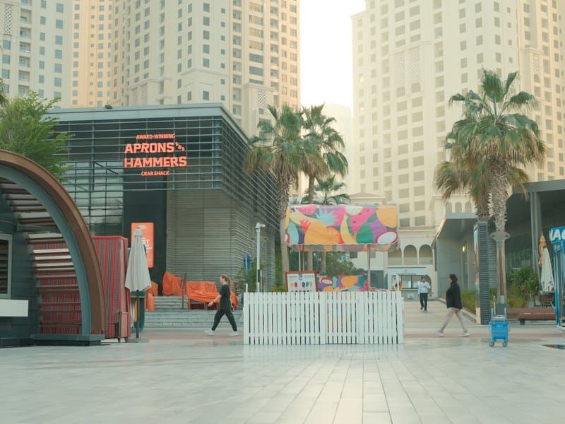 Urban scene featuring modern buildings, palm trees, and colorful art installation in a plaza.