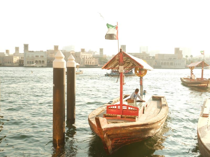 A traditional wooden boat docked near a waterfront with city buildings in the background under bright sunlight.