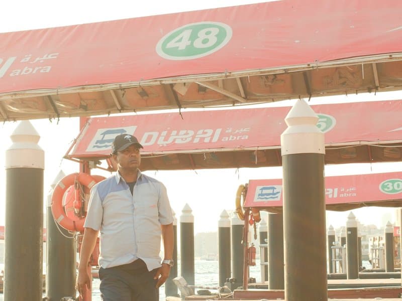 A man walking along a dock with red canopies and boat numbers in the background, bathed in warm sunlight.