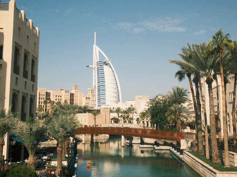 View of a modern building beside a canal, with palm trees and a bridge in the foreground.