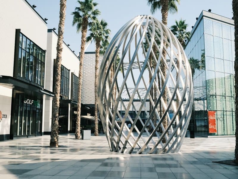 A modern metallic sculpture resembling an egg shape in an urban plaza surrounded by palm trees.