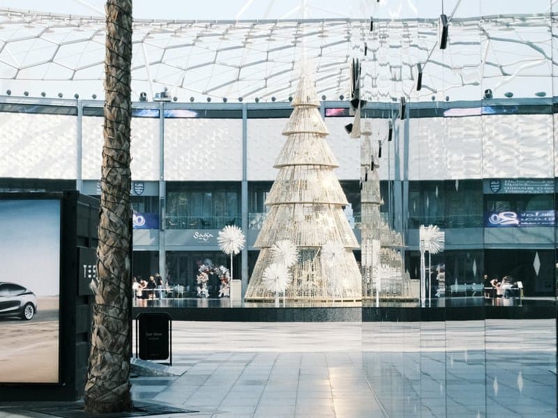 A modern shopping center featuring a large decorative Christmas tree in the center, surrounded by shoppers.