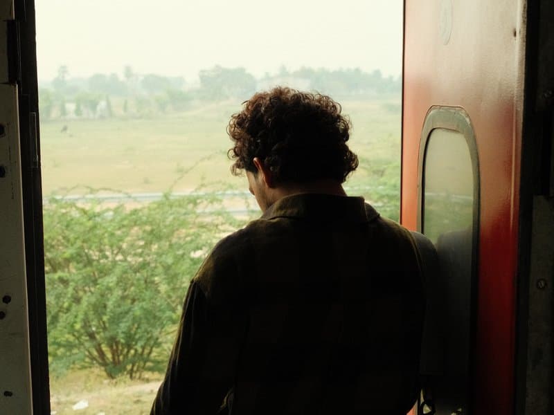 A person with curly hair stands at an open door, gazing out at a green landscape.