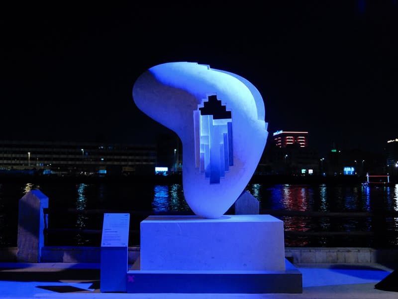 A modern sculpture illuminated in blue light at night, reflecting on water with city lights in the background.