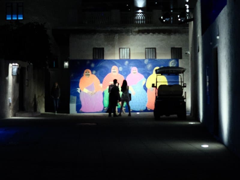 Silhouetted figures walking past a colorful mural in a dimly lit alley.