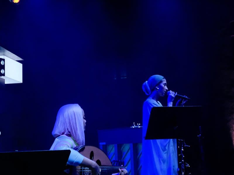 A singer performing on stage with blue lighting, accompanied by a musician playing an instrument.
