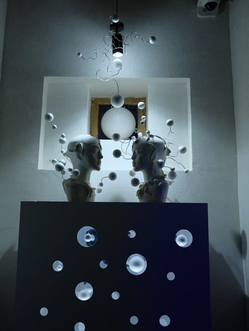 Two white sculptures of heads facing each other, surrounded by floating spheres and soft lighting.