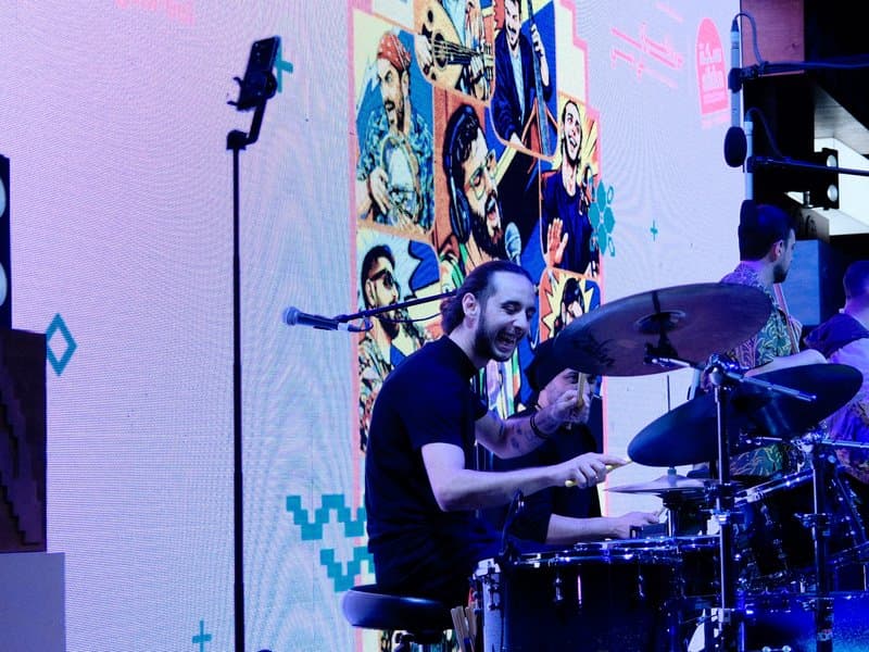 A drummer performing on stage with a colorful backdrop featuring animated characters.