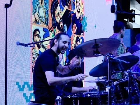 A drummer energetically playing on stage with colorful visuals in the background.