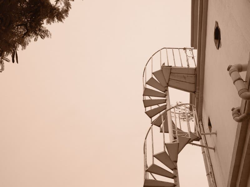 A spiral staircase ascending alongside a building, with a tree partially visible in the frame.