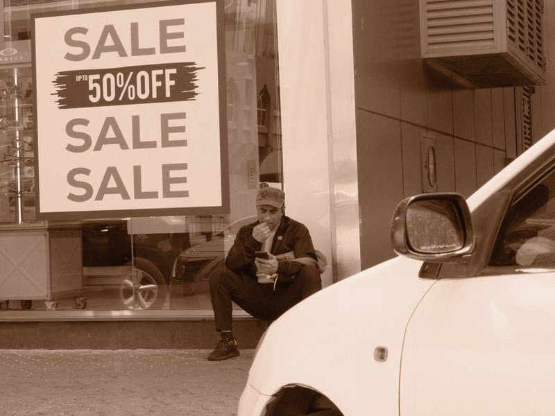 A man sitting outside a store with a large sale sign, a white car in the foreground.