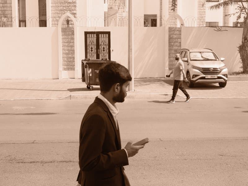 A man in a suit stands on the street looking at his phone, with a blurred pedestrian and cars in the background.