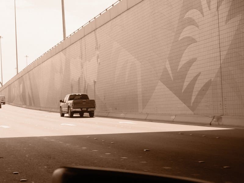 A pickup truck driving on a highway beside a mural-covered wall under a bright sky.
