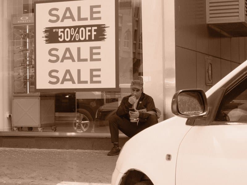 A man sitting outside a store with a sale sign, eating food, with a car in the foreground.