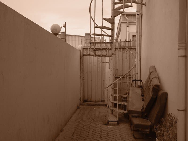 Narrow alleyway with a spiral staircase and two chairs against a wall.