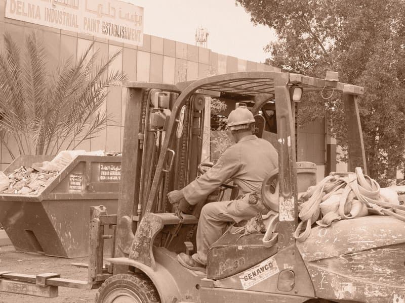 A worker operating a forklift in front of a building, surrounded by debris and greenery.