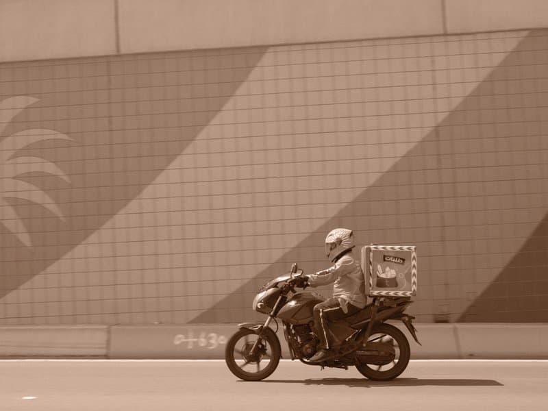 A motorcycle courier rides past a colorful mural with palm tree patterns on a city street.