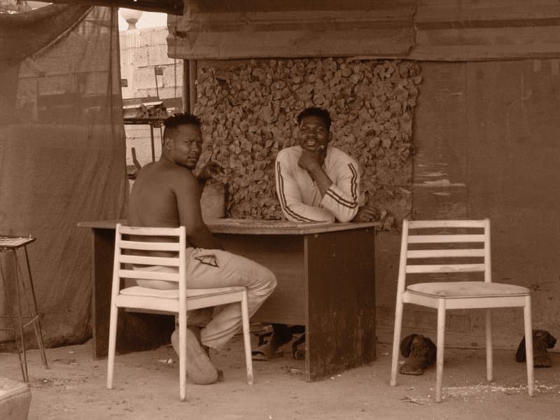 Two men seated at a table in a makeshift shelter, engaged in conversation.