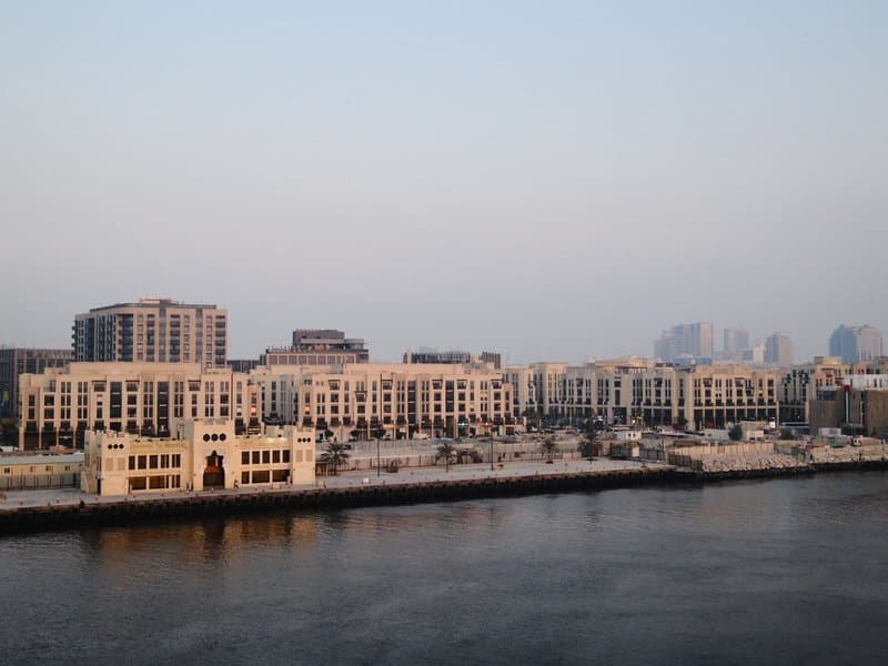 A waterfront view of modern buildings along a river at dusk, with soft light reflecting on the water.