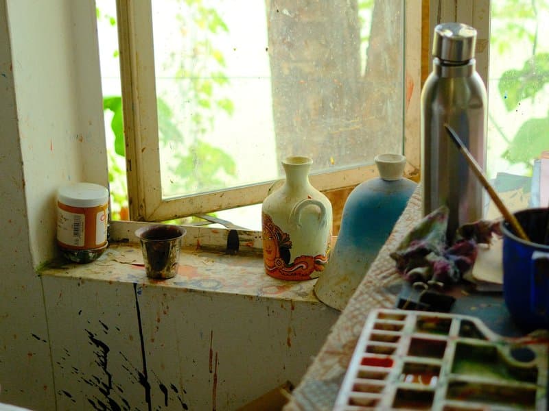 A sunlit windowsill with various jars, a ceramic vase, and a watercolor palette.