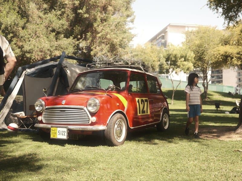 A vintage red car with racing stripes parked on green grass near a tent, with a child walking nearby.