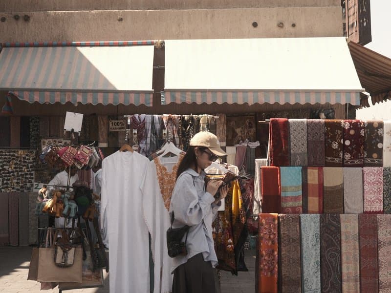 A woman holding a drink stands in front of a vibrant market stall filled with textiles and clothing.