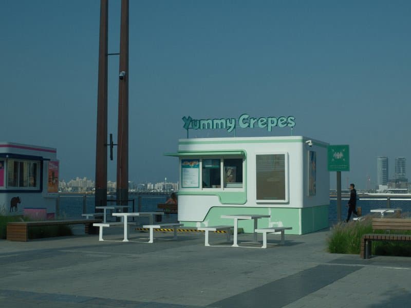 A pastel green food kiosk named 'Minty Grapes' by the waterfront with benches and a clear blue sky.