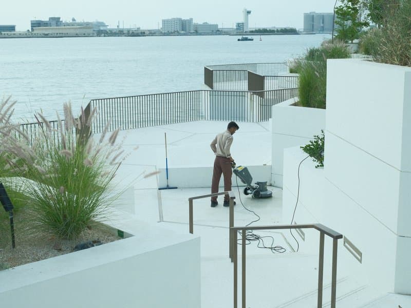 A person cleaning a white outdoor staircase near a waterfront with buildings in the background.
