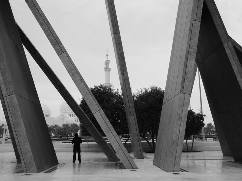 A person stands between large metallic structures with trees and a tower in the background on a cloudy day.