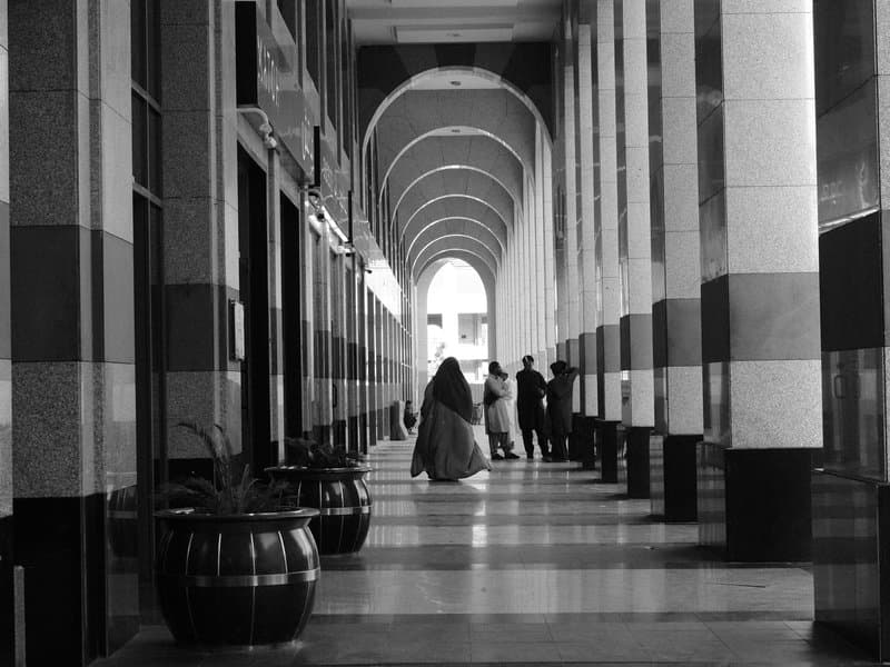 A black and white photo of a corridor with arches, featuring a person in a long coat and two figures in the distance.