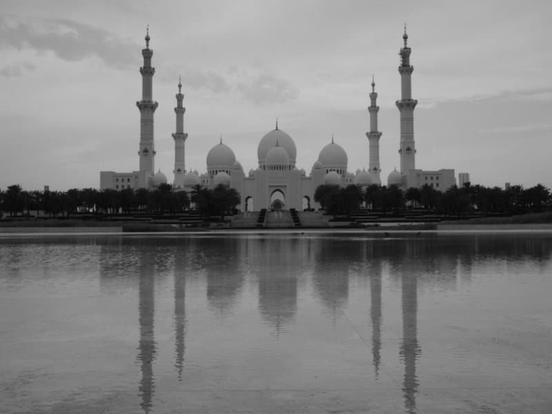 Black and white image of a grand mosque with multiple minarets reflected in a calm water surface.