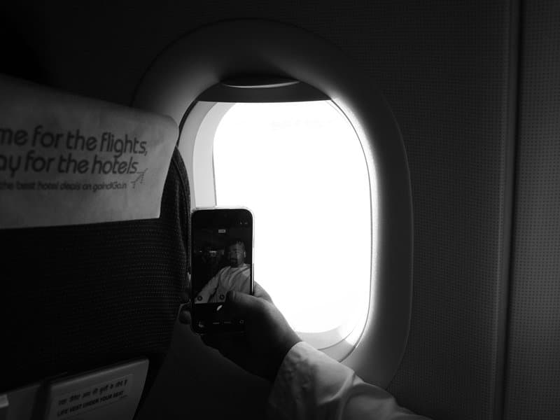 A hand holding a smartphone near an airplane window, with a bright white view outside.
