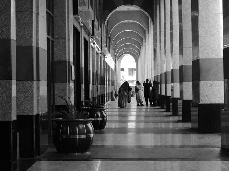 A monochrome image of a long corridor with arches, featuring people in conversation and decorative planters.