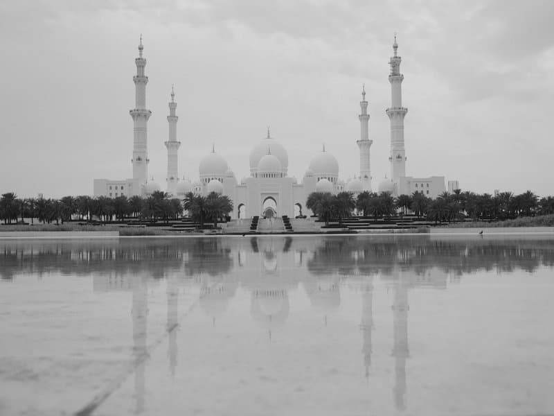A grand mosque with multiple domes and minarets reflected in a water surface under a cloudy sky.