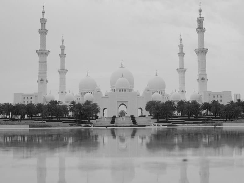 A grand mosque with multiple minarets reflected in calm water under a cloudy sky.