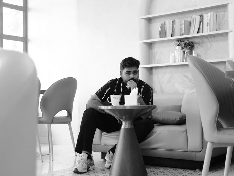A man sitting on a couch in a modern indoor setting, sipping from a cup with a table nearby.