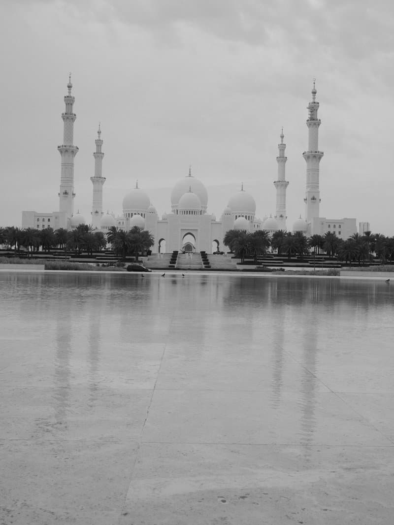 A grand white mosque with multiple minarets reflected in a calm water surface under a cloudy sky.