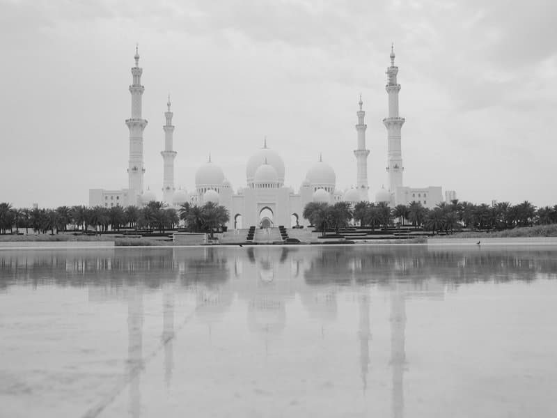 A grand mosque with multiple minarets reflected in a calm water surface under a cloudy sky.