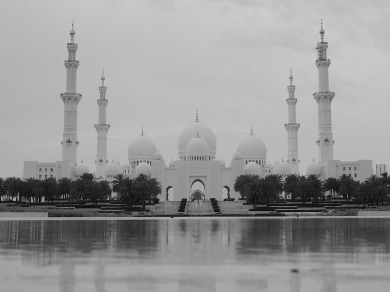 A grand mosque with multiple minarets and domes reflected in a calm body of water under a cloudy sky.