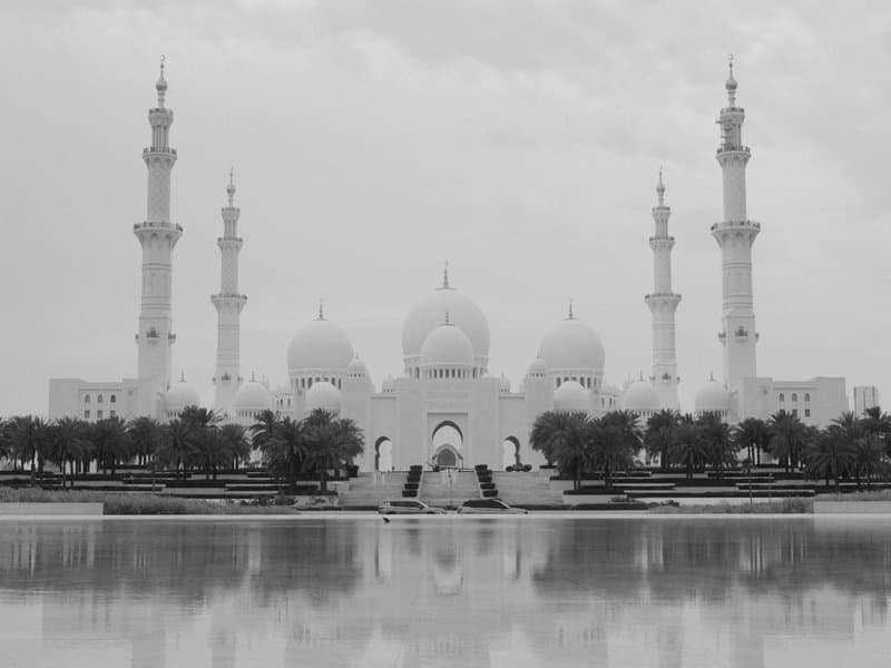 A grand mosque with multiple minarets reflected in a calm water body under a cloudy sky.