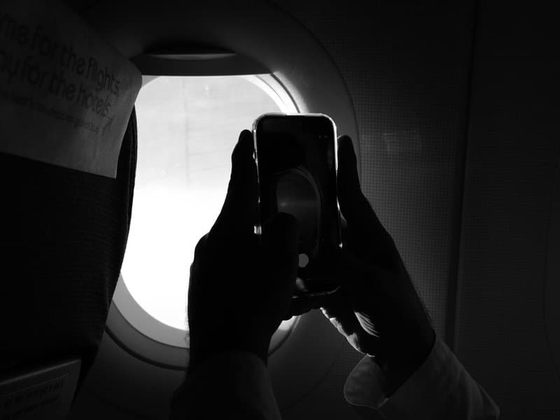 Silhouette of hands holding a smartphone near an airplane window with bright light outside.