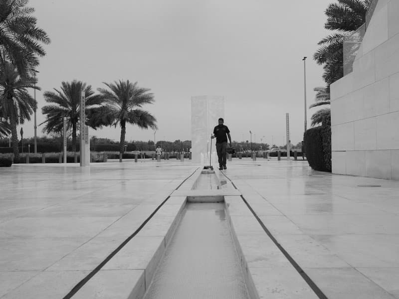 A lone figure walking along a reflective pathway in a modern outdoor space with palm trees.